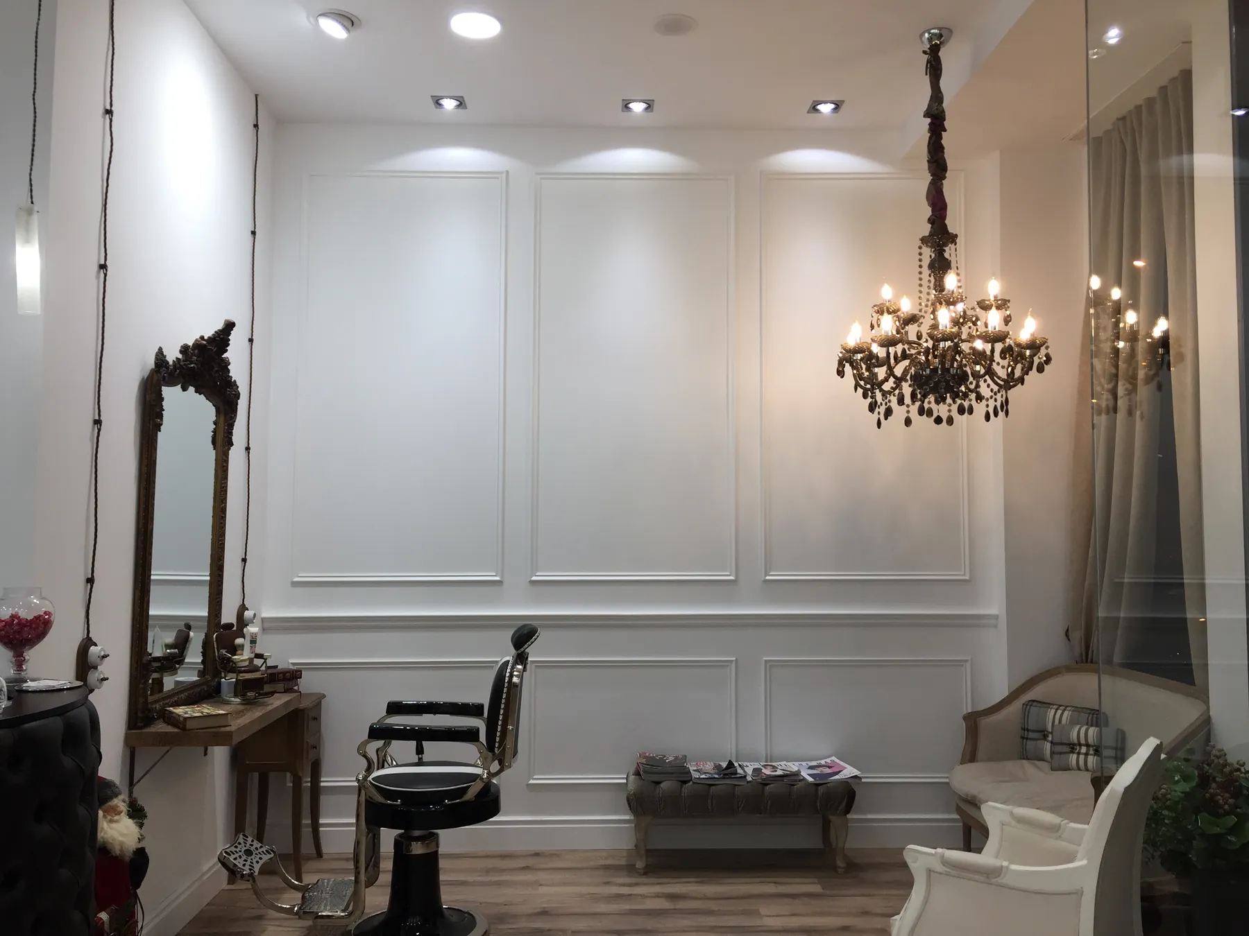 Interior de la peluquería con pared de molduras blancas, espejo ornamental y lámpara de araña.