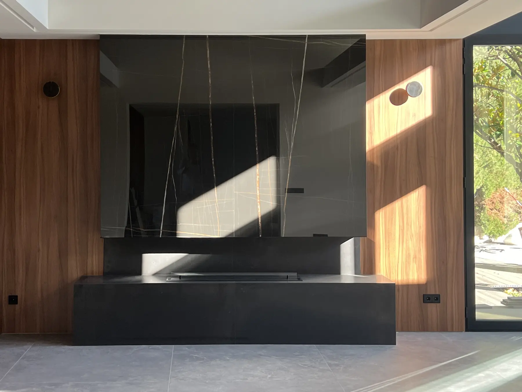 Panel oscuro veteado sobre un mueble bajo, entre superficies de madera y un ventanal.