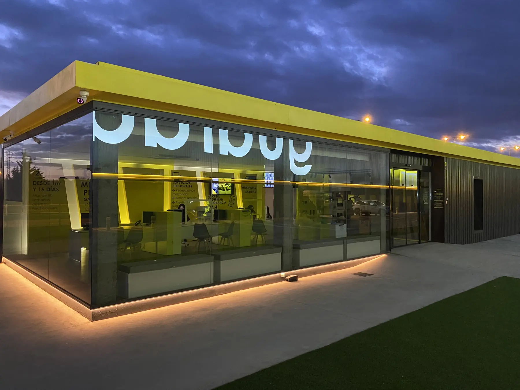 Fachada acristalada de Guardatodo al anochecer con remate amarillo e iluminación perimetral.