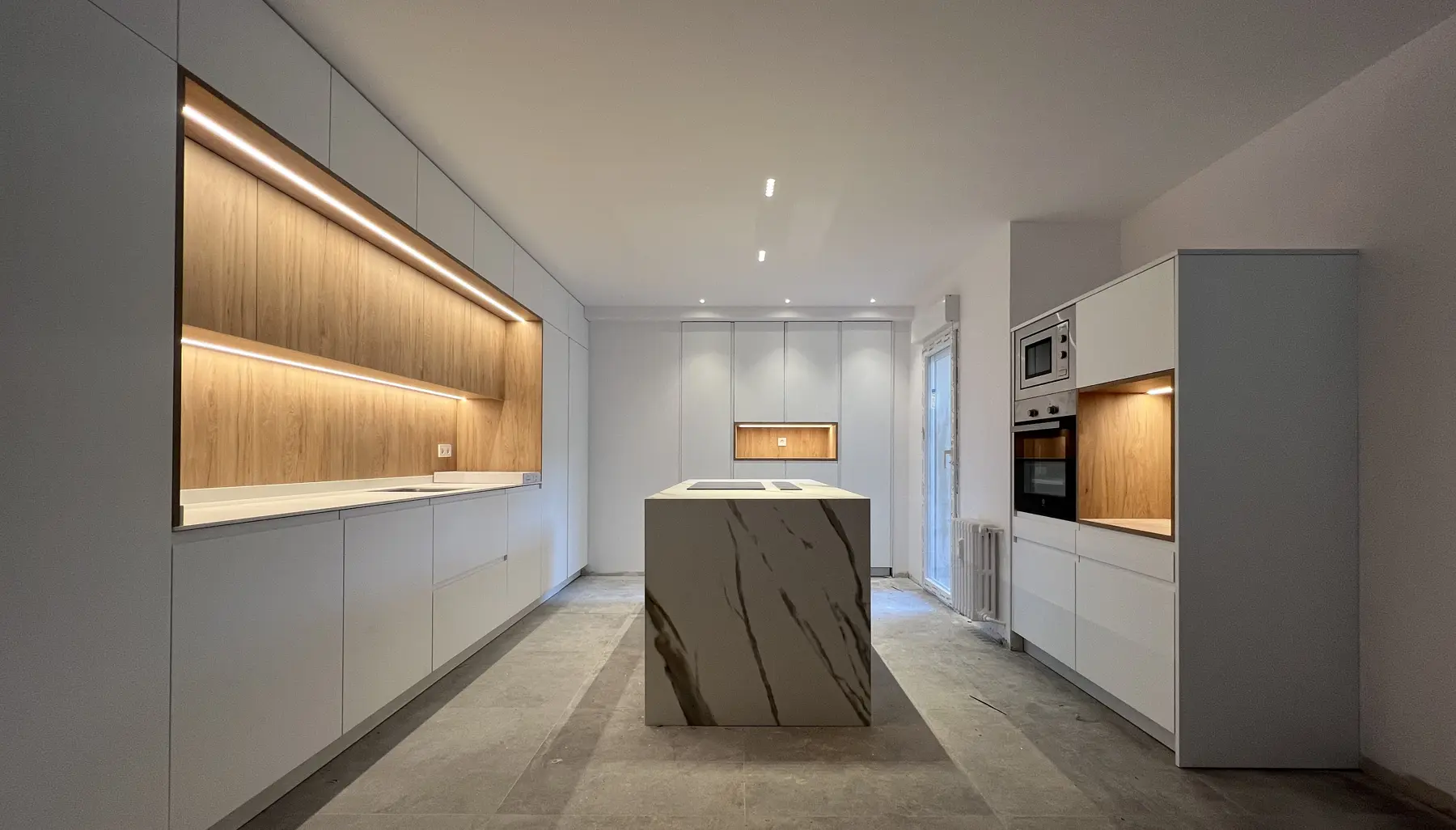 Vista alargada de la cocina con isla veteada, muebles claros y estantes iluminados.