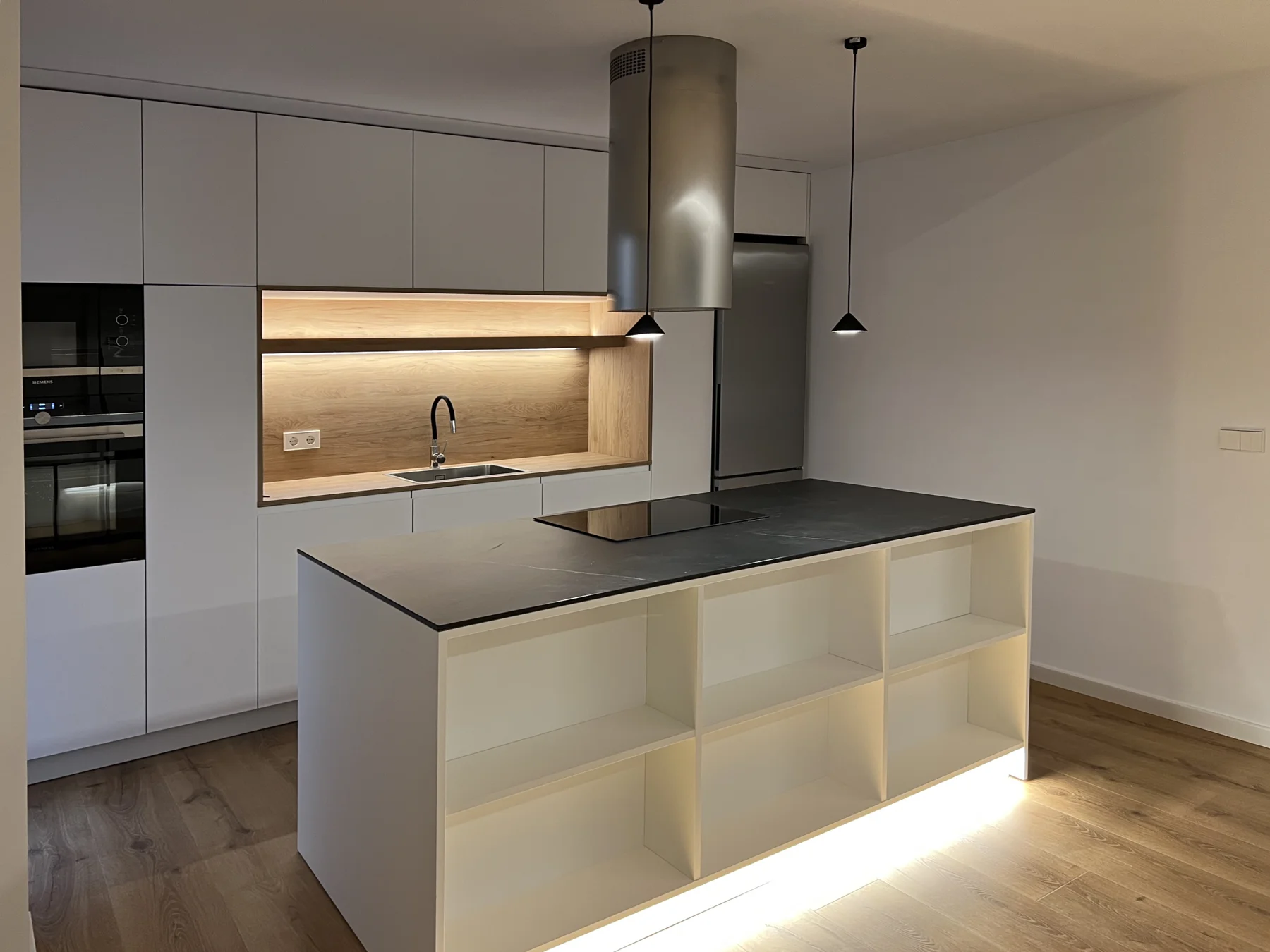 Isla de cocina con encimera oscura y estantes blancos iluminados en su frente.