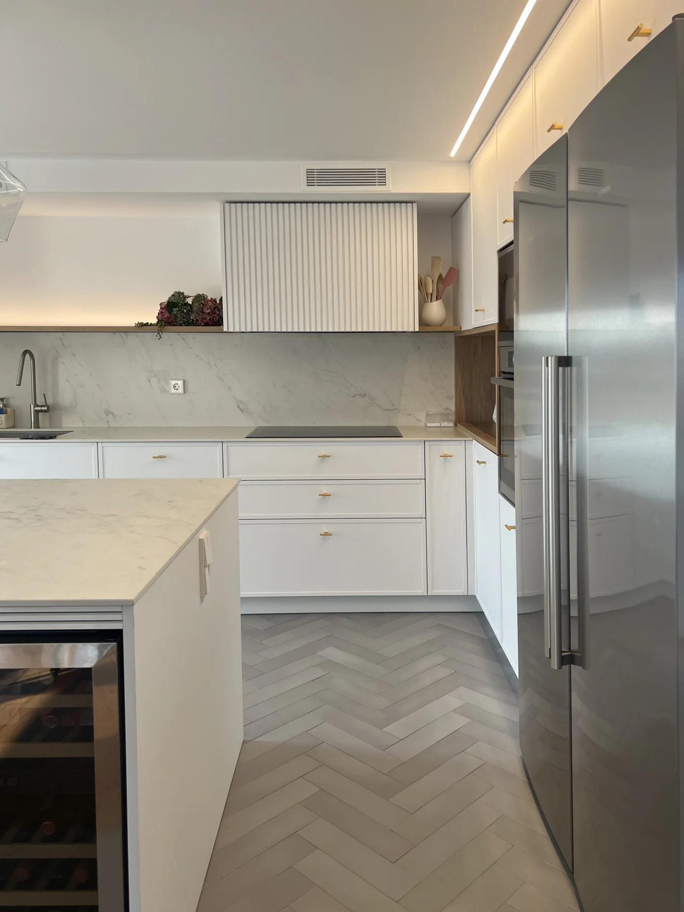 Cocina blanca con isla central, frigorífico metálico y pavimento en espiga.