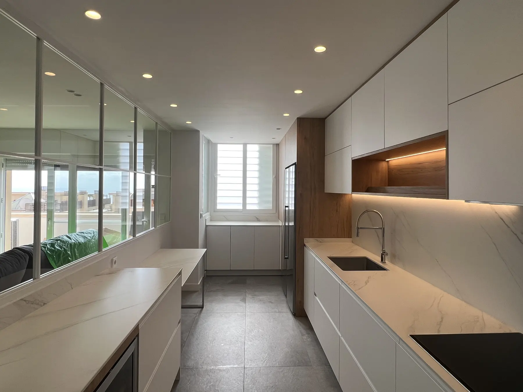 Cocina alargada con muebles blancos, detalles de madera y luz natural al fondo.